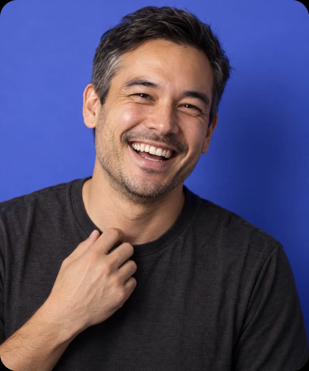 A man laughing with his hand at his collar in front of an indigo wall