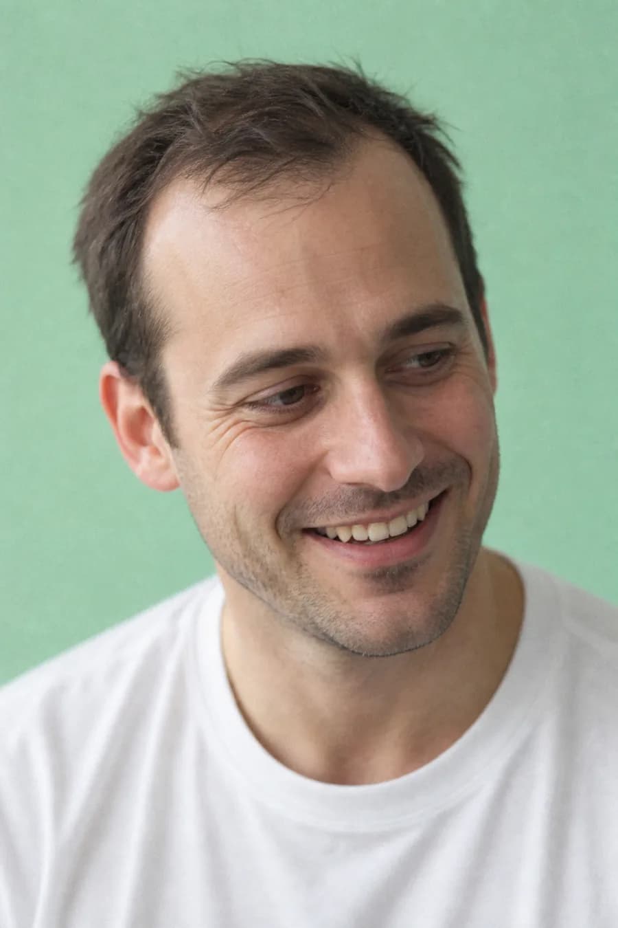 A man with a receding hairline laughing and looking off to the side in front of a mint green wall